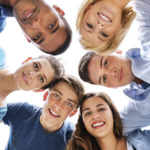 Six adolescent boys and girls stand in a circle, looking down at the camera and smiling.